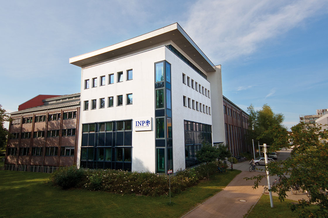 Außenansicht eines modernen Institutsgebäudes mit INP-Logo auf dem Campus in Greifswald, umgeben von Grünflächen und Wegen.