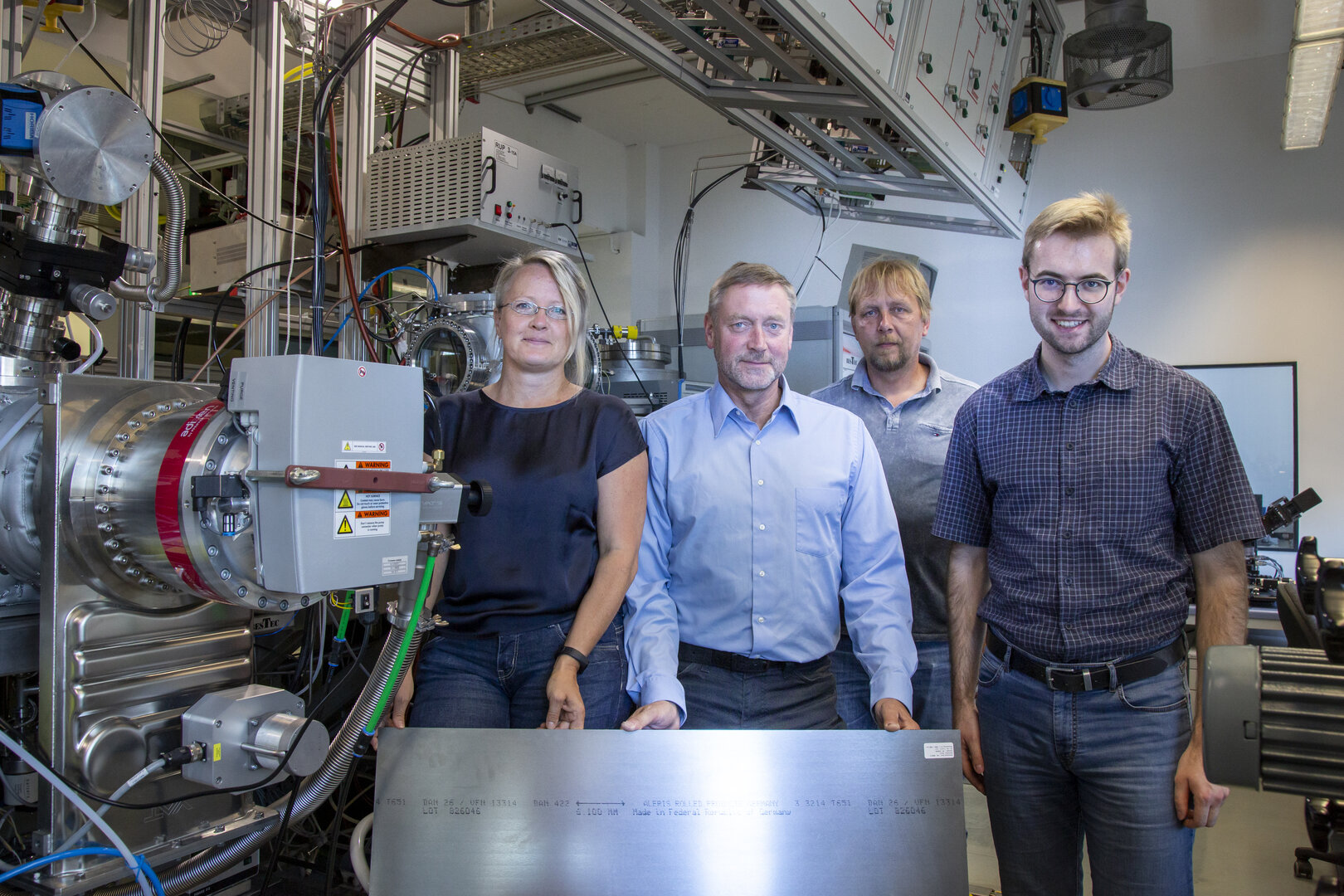 Dr. Angela Kruth, Prof. Klaus-Dieter Weltmann, Uwe Lindemann und Dr. Marcel Wetegrove stehen in einem Labor an einer Beschichtungsanlage und halten eine Stahlplatte für die Wasserstoffforschung.