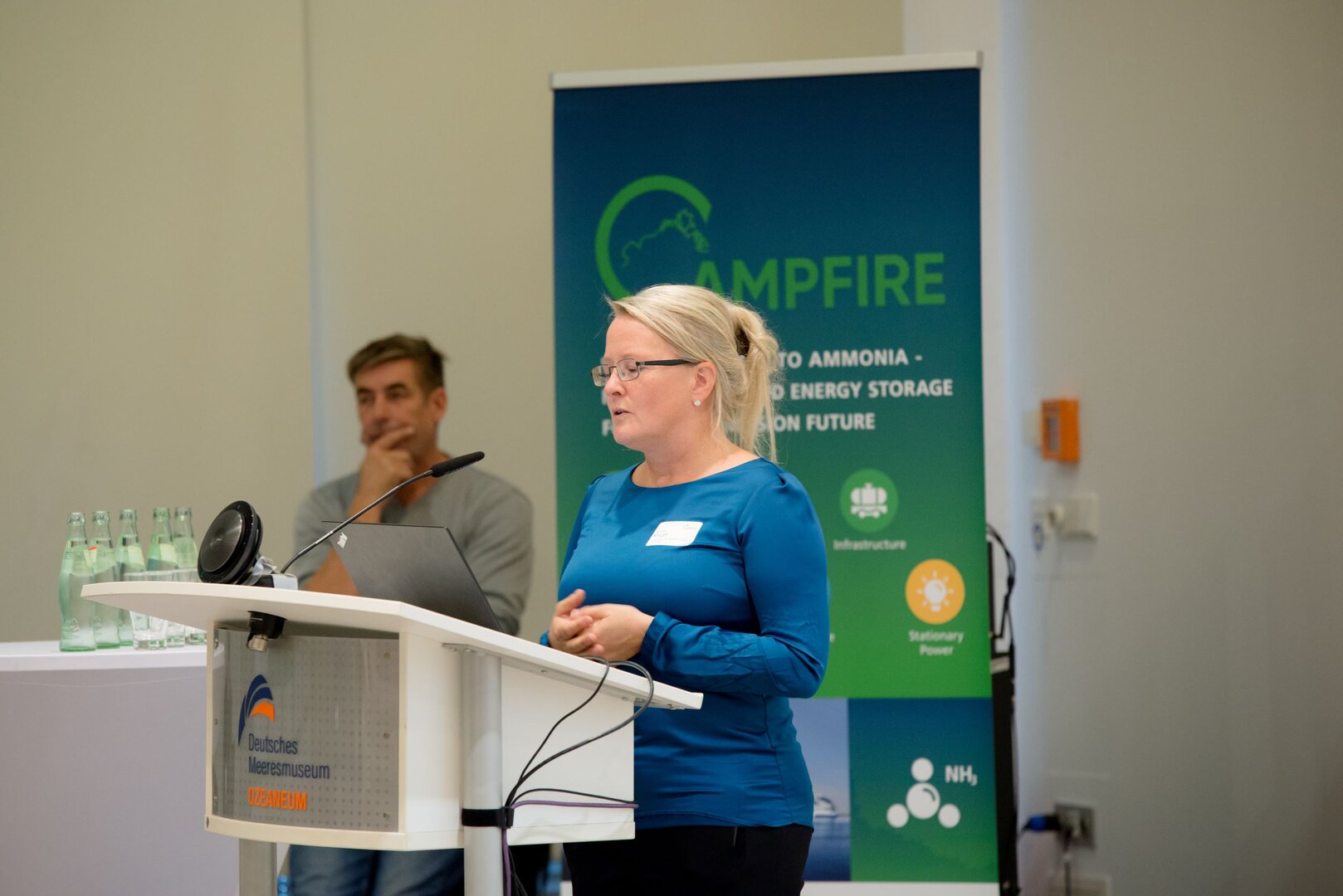 Eine Frau hält einen Vortrag an einem Rednerpult in einem Konferenzraum; im Hintergrund ist ein CAMPFIRE-Banner zu sehen, daneben sitzt eine weitere Person.