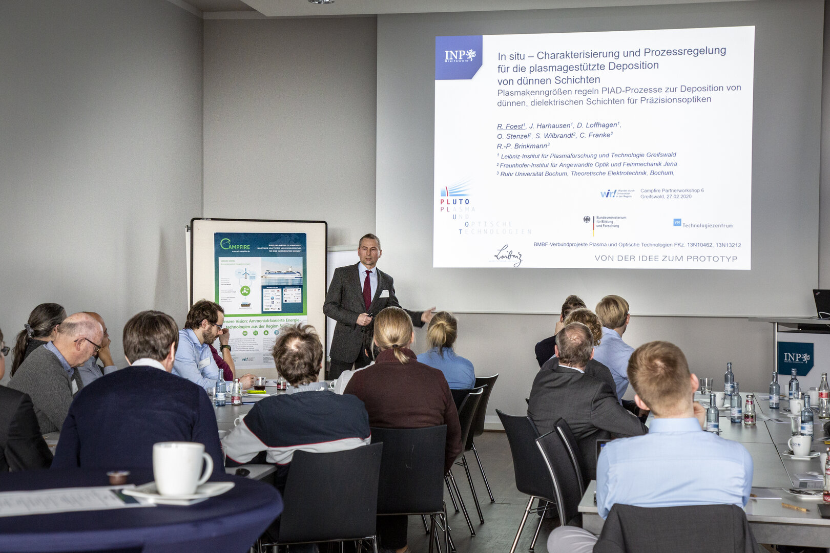 Eine Person hält einen Fachvortrag zu plasmagestützten Dünnschichtprozessen vor einer sitzenden Workshop-Gruppe; im Hintergrund sind Präsentationsfolie und CAMPFIRE-Poster zu sehen.