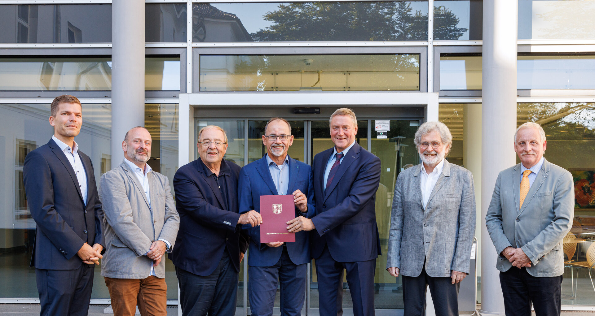 Minister Dr. Wolfgang Blank presents a funding notice to Prof. Klaus-Dieter Weltmann in front of the KDK building, accompanied by Prof. Dr. med. Wolfgang Motz and other participants. 