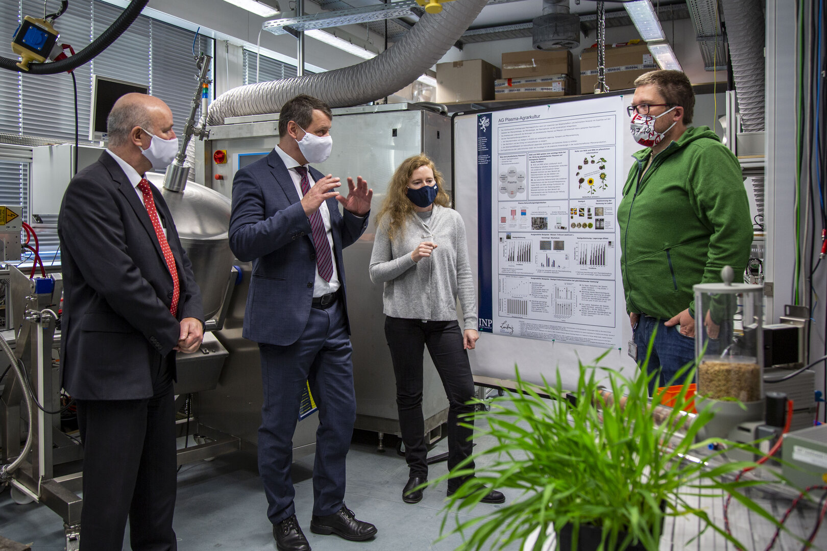 Michael Sack und Egbert Liskow besuchen ein INP-Labor, in dem Henrike Brust und Stefan Horn an einem Poster zu Plasmaanwendungen in der Agrarkultur erläutern; alle tragen Masken.
