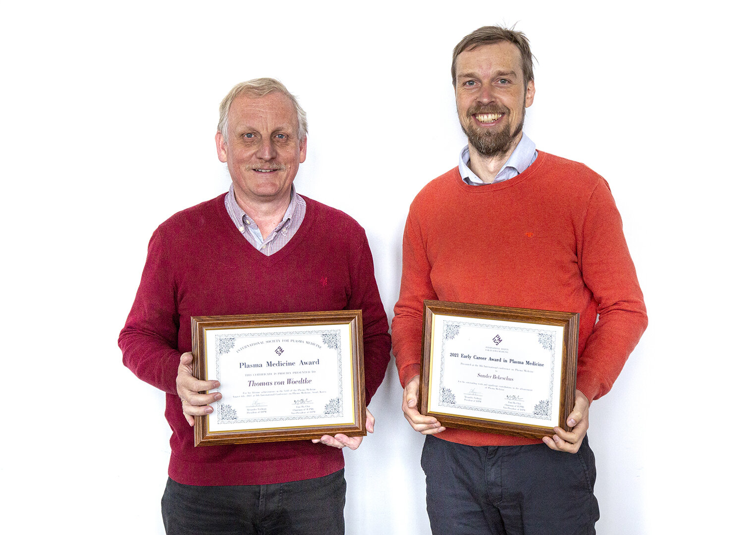 Two individuals hold framed certificates from the International Society for Plasma Medicine against a neutral background.