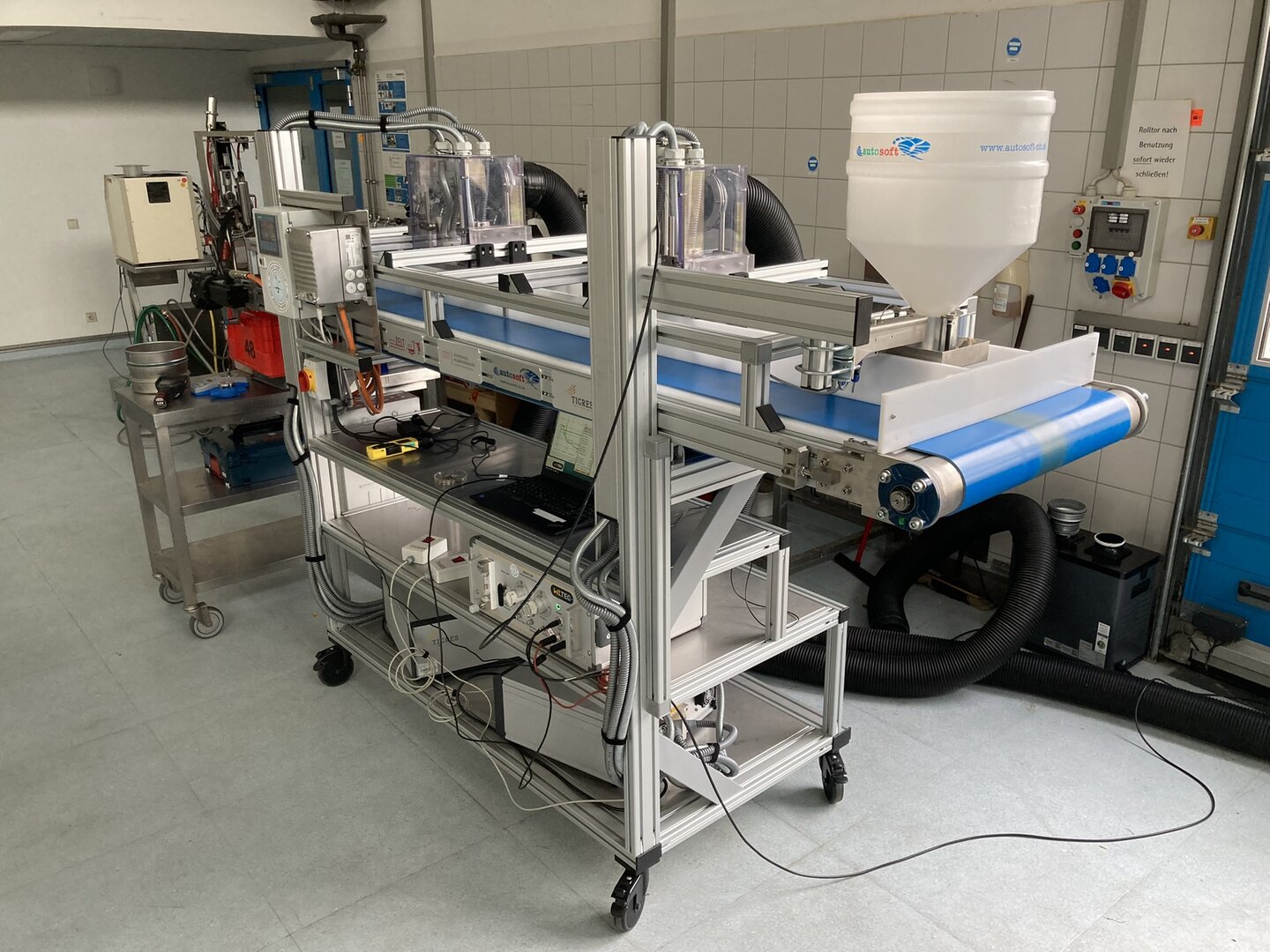 Versuchsaufbau mit modifiziertem Förderband zur plasmabasierten Behandlung von Schüttgut; Anlage mit Steuertechnik, Förderband, Behälter und Messtechnik in einem Laborraum.