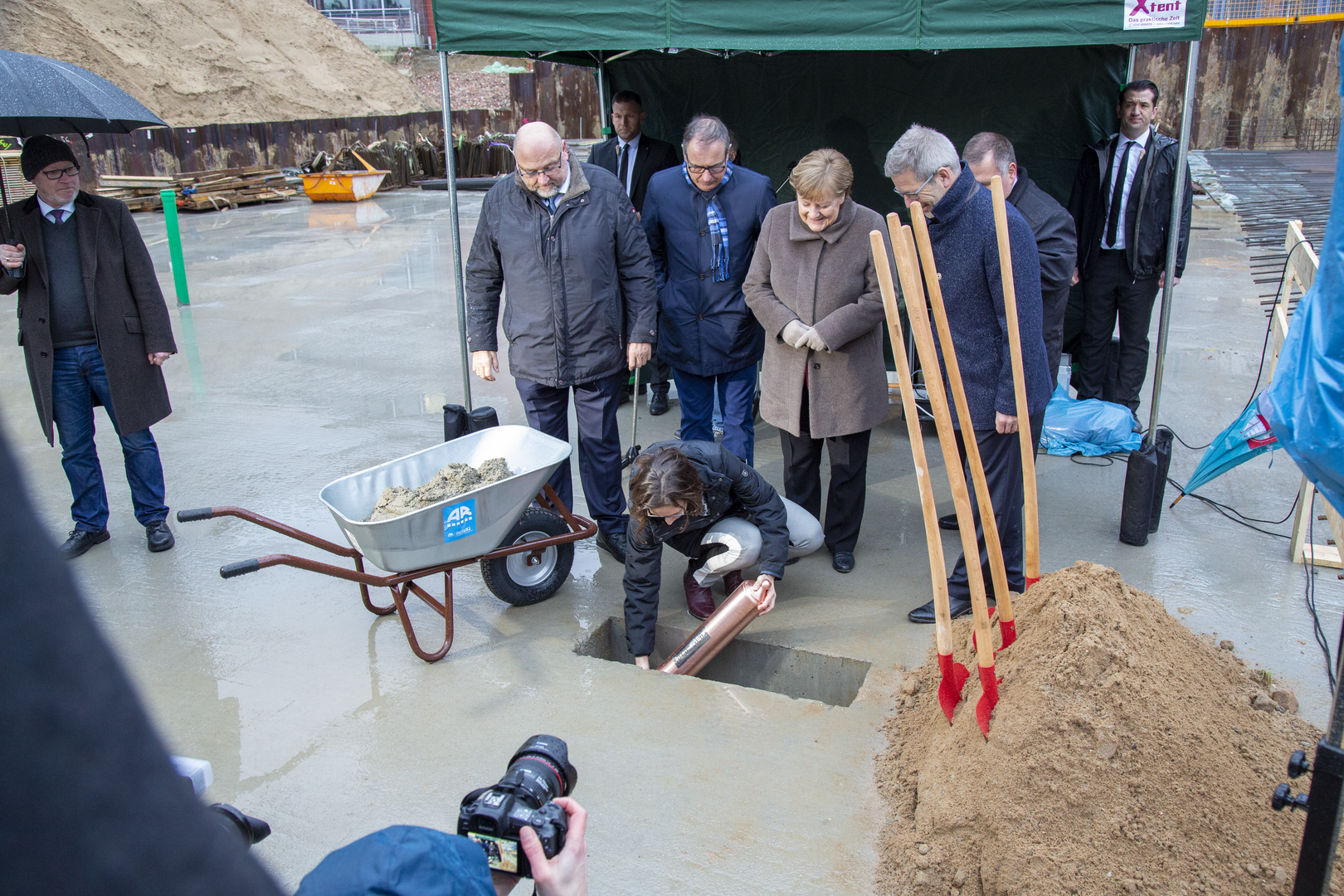 Mehrere hochrangige Vertreter aus Politik und Verwaltung begleiten eine symbolische Grundsteinlegung, bei der eine Person eine Metallkapsel in eine vorbereitete Öffnung im Beton einsetzt.